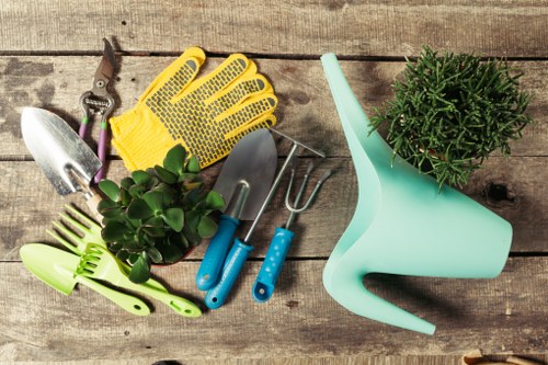 This image displays a collection of gardening tools and accessories arranged on a weathered wooden outdoor surface, likely part of a garden or yard. Prominent items include a pair of yellow and black gardening gloves, a small handheld trowel, a larger trowel, and a hand rake with blue handles. There is also a green watering can, and a small potted green shrub with dense foliage positioned near the watering can, suggesting plant care or gardening activity. The background and surface feature aged, textured wood, providing a rustic appearance suitable for outdoor gardening environments. The arrangement suggests preparation for gardening tasks such as planting, weeding, or lawn maintenance, aligning with services like lawn mowing and garden upkeep offered by Lawn Mowing Knightsbridge. The image's natural color tones and outdoor setting relate to gardening services provided in the Knightsbridge area, known for stylish outdoor landscapes and well-maintained gardens. Light appears natural, indicating an outdoor environment with soft shadows, suitable for outdoor gardening work in various weather conditions.